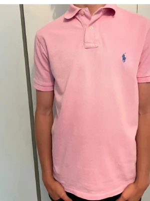 Rosa pikétröja från Polo Ralph Lauren - Snygg rosa pikétröja från Polo Ralph Lauren med blå logga på bröstet. Modellen är custom slim fit och har klassisk krage med knappar. Perfekt för en stilren och avslappnad look.