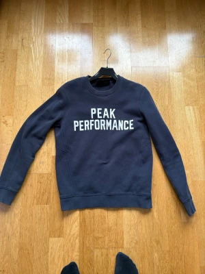 Mörkblå Peak Performance sweatshirt - Mörkblå sweatshirt från Peak Performance med vit logotyptext på bröstet. Klassisk rund halsringning och ribbade muddar vid ärmslut och nederkant. Perfekt för en avslappnad och sportig stil.