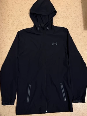 Svart vindjacka från Under Armour - Svart vindjacka med luva från Under Armour. Jackan har hel dragkedja framtill, två snedställda fickor med dragkedja och justerbar dragsko i luvan. Logga på bröstet och muddar med Under Armour-tryck. Perfekt för blåsiga dagar.