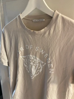 Beige t-shirt från Tiger of Sweden - Snygg beige t-shirt från Tiger of Sweden med vitt tryck framtill och rund halsringning. T-shirten har en klassisk passform och är tillverkad i mjuk bomull, perfekt för en stilren och avslappnad look.