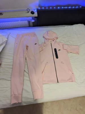 Rosa Nike tech set - Säljer ett rosa Nike tech set från Nike med matchande byxor och hoodie. Hoodien har dragkedja, fickor och svart Nike-logga på bröstet. Byxorna har resår i midjan och muddar vid bensluten. Perfekt för chill dagar hemma.
