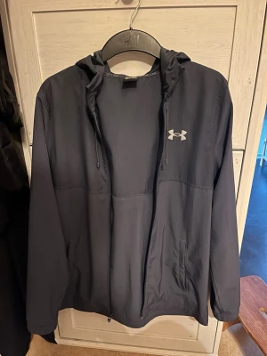 Mörkblå vindjacka från Under Armour - Snygg mörkblå vindjacka från Under Armour med huva och dragkedja framtill. Jackan har två sidofickor, justerbar huva med dragsko och logga på bröstet samt stor text på ryggen. Tillverkad i lätt och vindtätt material, perfekt för blåsiga dagar.