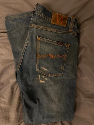 Blå Nudie Jeans med slitningar - Säljer ett par blå jeans från Nudie Jeans med klassisk femficksdesign och orangea sömmar. Jeansen har slitningar och detaljer på bakfickorna med Nudie-signaturen. Materialet är denim w29 L32