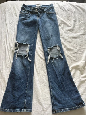 Blå slitna bootcut jeans med hål - Säljer ett par blå bootcut jeans med stora slitna hål på båda knäna. Jeansen har klassisk femficksdesign och är gjorda i denim. Perfekt för dig som gillar en edgy och avslappnad stil.