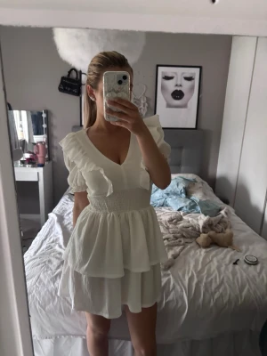 Vit volangklänning med v-ringning - Säljer en vit kortklänning med v-ringning och volangdetaljer på axlarna. Klänningen har smockad midja och dubbla lager i kjolen som ger en luftig och feminin look. Perfekt för sommarens alla tillfällen!