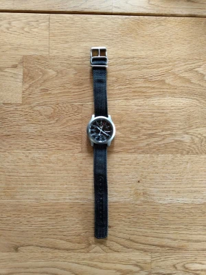 Seiko klocka med svart textilband - Klassisk Seiko armbandsklocka med genomskinlig baksida där urverket syns. Klockan har ett svart textilband och silverfärgat boett. Perfekt för dig som gillar stilrena accessoarer med mekaniskt urverk.
