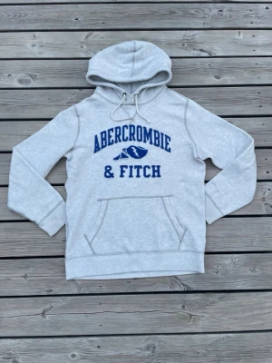 Abercrombie & Fitch hoodie! - Snygg ljusgrå hoodie från Abercrombie & Fitch med blå logga och text framtill. Klassisk känguruficka och dragsko i huvan.  Den är lite knopprig på texten annars är det inget fel på den, fråga vid fundering!😁😇
