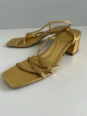 ZARA guldiga sandaler med flätade remmar, storlek 40 - Snygga sandaler från ZARA i storlek 40 i metallic guld med fyrkantig tå och flätade remmar. Skon har en stabil blockklack och öppen design som ger en trendig look. Perfekta för att lyfta vilken outfit som helst med sin glansiga finish och moderna form. Mycket gott skick!!