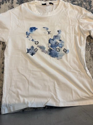 Vit t-shirt med blått tryck - Säljer en vit t-shirt med blått och marinblått vattenfärgstryck med snygga tecken på bröstet. T-shirten har rund hals och korta ärmar. Perfekt för dig som gillar exklusiva streetwear-plagg.