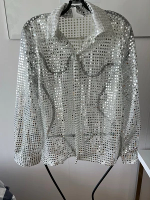 Glittrig discotröja i silver - Säljer en glittrig discotröja i silver med långa ärmar och stora paljetter. Skjortan har klassisk krage och är tillverkad i 100% polyester. Perfekt för maskerad, temafest eller cosplay med disco- eller 70-talsstil. Några paljetter har ramlat av. 