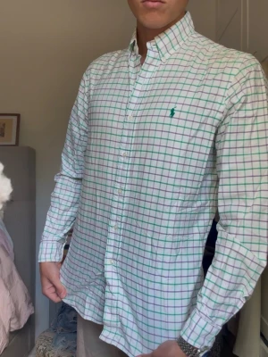 Rutig skjorta från Ralph Lauren - Snygg vit skjorta med grönt och rosa rutmönster från Ralph Lauren. Klassisk button-down krage och långärmad modell med grönt broderat logotyp på bröstet. Perfekt för en stilren och fräsch look. (Passform: jag på bilden är 190 lång och väger 80kg)