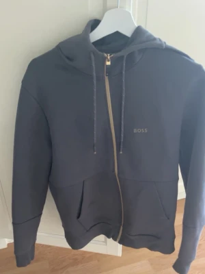 Mörkgrå hoodie från BOSS med dragkedja - Snygg mörkgrå hoodie från BOSS med dragkedja framtill, justerbar huva med snören och två stora fickor. Diskret BOSS-logga på bröstet. Perfekt för en avslappnad och stilren look.