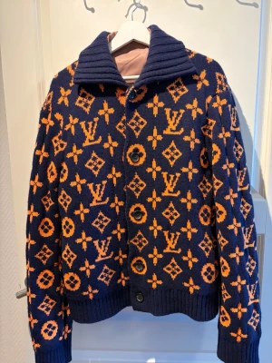 Mörkblå Louis Vuitton stickad kofta - Säljer en mörkblå stickad kofta från Louis Vuitton med orange monogrammönster över hela plagget. Koftan har stora knappar framtill, bred krage och ribbstickade muddar vid ärmslut och nederkant. Perfekt för dig som vill sticka ut med lyxig design. Box + digital kvitto 