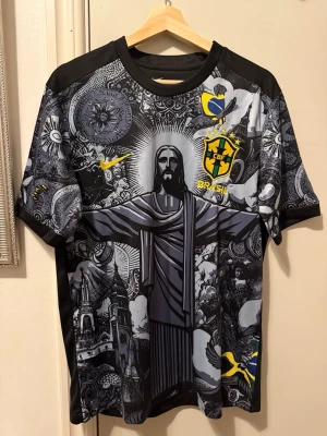 Brasil Nike fotbollströja med tryck - Unik svart och grå Brasilien fotbollströja från Nike med detaljerat grafiskt tryck av Kristusstatyn och brasilianska symboler. Tröjan har CBF-emblem, gula detaljer och korta ärmar. Perfekt för dig som vill sticka ut på planen eller läktaren.