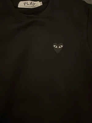 Svart sweatshirt Comme des Garçons Play - Svart sweatshirt från Comme des Garçons Play med det ikoniska hjärta med ögon på bröstet. Tröjan har rund halsringning, långa ärmar och ribbade muddar. Perfekt för en stilren och trendig look.
