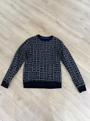 Svart sweatshirt med HUGO-logga -  Svart sweatshirt från HUGO med grått logomönster över hela tröjan. Rund halsringning och ribbade muddar vid ärmslut och nederkant. Perfekt för dig som gillar streetstyle och vill sticka ut med en snygg logga.