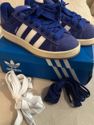 Blå Adidas Campus sneakers - Snygga blå Adidas Campus sneakers i mocka med klassiska vita ränder på sidorna och vit sula. Skorna har blå skosnören och kommer även med extra vita och blå skosnören. Perfekt för dig som gillar retrostil och vill sticka ut med färg. Använda ett fåtal gånger.