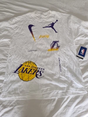 Nike Los Angeles Lakers T-shirt M - Snygg vit Nike T-shirt med Los Angeles Lakers tryck i gult och lila. T-shirten har flera loggor, inklusive NBA, Nike och Jordan, samt stor Lakers-boll på framsidan. Loose fit-modell i mjuk bomull, perfekt för dig som gillar basket och streetwear.