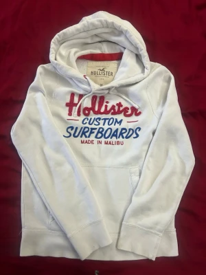  hoodie Hollister  - Vit hoodie från Hollister med broderat och tryckt text i rött och blått på bröstet: 'Hollister Custom Surfboards Made in Malibu'. Klassisk känguruficka och dragsko i huvan. Mjuk bomullsmix och avslappnad passform.
