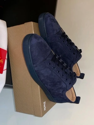 Mörkblå sneakers från Louboutin - Snygga mörkblå sneakers från Christian Louboutin i mocka med matchande mörkblå sula och svarta skosnören. Diskret broderad logga på hälen och klassisk låg modell. Perfekta för dig som vill ha en stilren och exklusiv look.