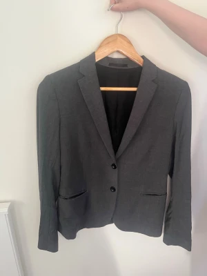 Mörkgrå klassisk kavaj - Stilren mörkgrå kavaj med två knappar framtill och två fickor. Kavajen har smal passform, klassisk krage och helsvart foder. Perfekt till en dressad look och enkel att matcha med olika plagg. Materialet ser ut att vara en blandning av polyester och viskos.