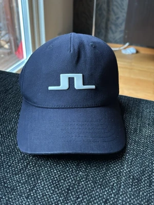 Marinblå keps från J.Lindeberg - Snygg marinblå keps från J.Lindeberg med böjd skärm och det klassiska JL-logotypen framtill i vitt. Kepsen är i flex-modell och har små ventilationshål upptill. Tillverkad i ett mjukt och flexibelt material som sitter skönt på huvudet.