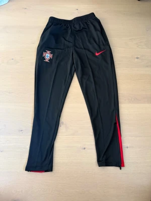 Portugal Nike träningsbyxor svart/röd - Snygga svarta träningsbyxor från Nike med Portugals landslagsmärke på ena benet och röd Nike-logga. Byxorna har röda detaljer vid benslut och är tillverkade i ett lätt och sportigt material med resår i midjan. Storleken är M, men passar S