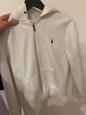 Vit hoodie från Polo Ralph Lauren - Snygg vit hoodie med dragkedja från Polo Ralph Lauren. Klassisk design med huva, känguruficka och den ikoniska broderade loggan på bröstet. Perfekt för en clean och stilren look. Tillverkad i mjukt bomullsmaterial som är skönt att ha på sig.
