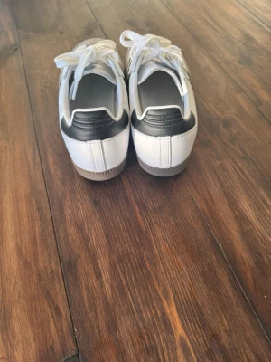 Adidas samba vita sneakers - Snygga Adidas Samba sneakers i vitt med svarta detaljer och klassiska tre ränder på sidorna. Ovandel i läder och mocka, vit sula och svart hälparti. Perfekta för en stilren och sportig look. Sköna snörskor med låg profil och retrovibbar.