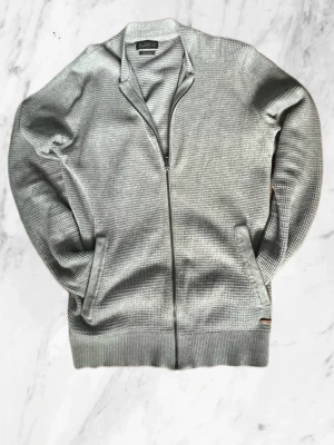 Grå ziptröja från Jack & Jones - Snygg grå ziptröja från Jack & Jones Originals med ribbad krage och muddar. Tröjan har två fickor framtill och är tillverkad i ett mjukt, stickat material. Perfekt för lager-på-lager och enkel att matcha med andra plagg.