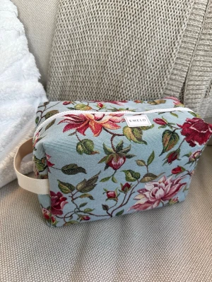 Blommig necessär från Emelo - Necessär i tyg från Emelo med ljusblå botten och blommor i rött, rosa och grönt. Rektangulär form med dragkedja upptill och ett praktiskt handtag i beige. Perfekt för smink eller småprylar. Innerfack finns. Mått ca 26x17x12cm