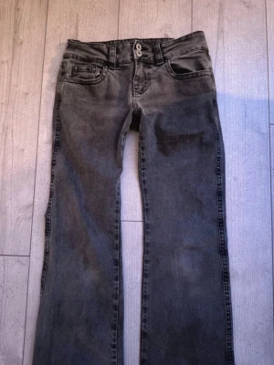 Grå bootcut jeans från Gina Tricot - Snygga grå bootcut jeans från Gina Tricot med klassisk femficksdesign och dubbla knappar i midjan. Jeansen har en lätt tvättad look och raka ben som går ut lite nertill. Jeansen har knappfickor på baksidan. Perfekta för en avslappnad och trendig stil.
