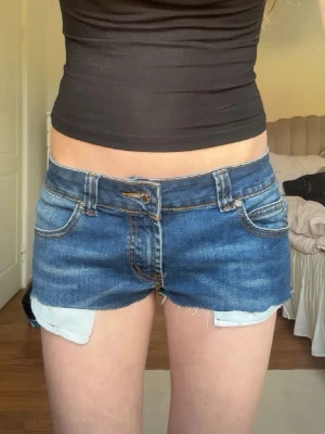 Blå jeansshorts från Nudie Jeans - Säljer ett par blå jeansshorts från Nudie Jeans med råa kanter och klassiska fickor både fram och bak. Shortsen har låg midja och är tillverkade i bomull. Shortsen har några små referenser som man ser på bild 3 och bild 4 