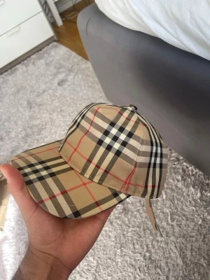 Beige Burberry keps med rutigt mönster - Säljer en klassisk beige keps från Burberry med det ikoniska rutiga mönstret i svart, vitt och rött. Kepsen har böjd skärm och justerbar rem bak. Tillverkad i bomull och polyester, perfekt för dig som vill ha en stilren accessoar med lyxig känsla.