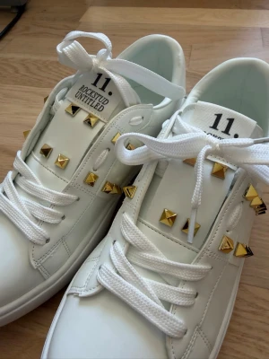 Vita sneakers med nitar - Snygga vita sneakers med coola guldfärgade pyramidnitar på sidorna och ovandelen. Skorna har klassisk låg modell, vita skosnören och en unik sula med rutmönster. Tillverkade i läder för en lyxig känsla.