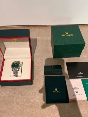 Role klocka med grön urtavla - Exklusiv Role klocka med grön urtavla och silverfärgat armband i metall. Klockan har en bezel med stenar runt urtavlan och klassisk länkdesign. Levereras med originalbox, certifikat och tillbehör. Perfekt för dig som vill sticka ut med stil.