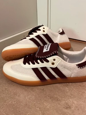 Adidas Wales Bonner vita sneakers - Snygga Adidas Wales Bonner sneakers i vitt med vinröda detaljer och klassiska tre ränder på sidan. Ovandelen är i mocka och läder, med brun gummisula och matchande vinröda skosnören. Tungan har Wales Bonner-logga och kontrastsömmar.
