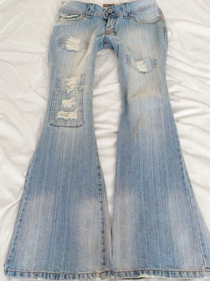 Ljusblå utsvängda jeans med slitningar - Säljer ett par ljusblå jeans med rejäl utsvängd passform och coola slitningar på låren och knäna. Jeansen har klassisk femficksdesign och låg midja. Perfekta för dig som gillar en retro och avslappnad stil.