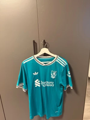 Turkos Liverpool matchtröja Adidas - Säljer en turkos Liverpool FC matchtröja från Adidas med vita detaljer och klubbmärke på bröstet. Tröjan har klassiska Adidas-ränder på axlarna och vit-svart krage. Tillverkad i lätt och ventilerande material, perfekt för fotboll eller träning.