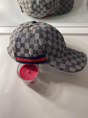  keps med GG-mönster - Snygg keps i beige och marinblått med klassiskt GG-monogram över hela kepsen. Kepsen har en böjd skärm och justerbar rem bak samt en röd och marinblå rand. Materialet är canvas med insida i tyg.
