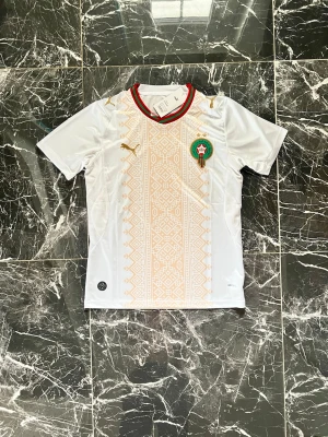 Marocko fotbollströja Puma vit - Vit Marocko fotbollströja från Puma med guldiga detaljer och traditionellt mönster framtill. Tröjan har rund hals med röd och grön kant samt Marockos landslagsemblem på bröstet. Perfekt för dig som vill visa ditt stöd på matchen.
