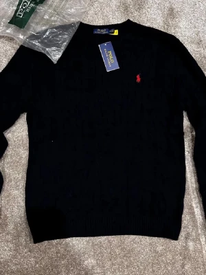 Svart kabelstickad tröja Polo Ralph Lauren - Snygg svart kabelstickad tröja från Polo Ralph Lauren med klassisk röd logga broderad på bröstet. Tröjan har rund halsringning och ribbade muddar vid ärmslut och nederkant. Perfekt för dig som gillar stilrena och tidlösa plagg.
