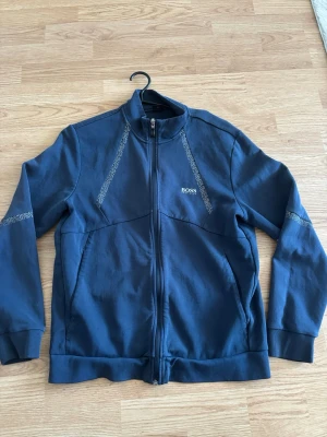 Mörkblå zip hoodie från BOSS - Snygg mörkblå hoodie från BOSS med hel dragkedja och diskreta mönstrade detaljer längs axlar och bröst. Tillverkad i mjukt material och har ribbade muddar vid ärmslut och nederkant. BOSS-logga på bröstet ger en clean och exklusiv look.