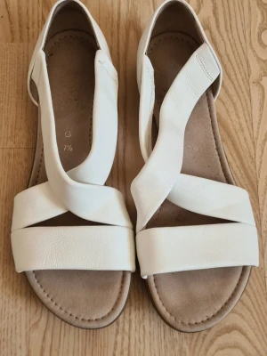 Vita sandaler med korsade remmar - Snygga vita sandaler med breda korsade remmar i mjukt konstläder. Sandalerna har öppen tå och en bekväm innersula i mockaimitation. Perfekta för sommaren och ger en stilren look till alla outfits.