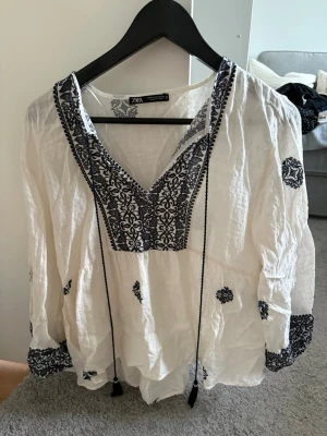 Vit blus från ZARA - Vit blus från ZARA med svart broderi längs v-ringningen, ärmslut och utspridda mönster. Långärmad modell med knytband framtill. Super fin och somrig blus!