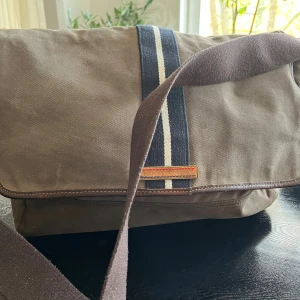 Brun axelväska Tommy Hilfiger - Snygg brun axelväska från Tommy Hilfiger i bomullscanvas med läderdetaljer. Väskan har ett brett axelband, blå och vit rand på locket samt randigt innerfoder med logga. Perfekt för dig som vill ha en stilren och praktisk väska med coola detaljer.