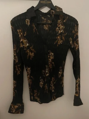 Plisserad svart blus med mönster - Snygg svart plisserad blus med bruna och beige mönster. Blusen har lång ärm, krage och knäppning framtill! Köpt på vinted och använder den tyvärr inte, uppskattar att den är en storlek M❤️
