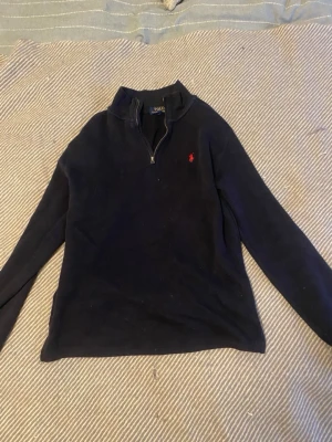 Mörkblå halvzip tröja Polo Ralph Lauren - Mörkblå långärmad tröja från Polo Ralph Lauren  halv dragkedja och hög krage. Tröjan har en röd broderad logga på bröstet och är tillverkad i mjukt stickat material. Perfekt för dig som gillar klassisk och stilren design.