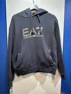 Svart hoodie från EA7 Emporio Armani - Snygg svart hoodie från EA7 Emporio Armani med stor logga i vitt och guld på bröstet. Hoodien har känguruficka, justerbar huva med dragsko och ribbade muddar. Perfekt för en avslappnad och trendig stil.