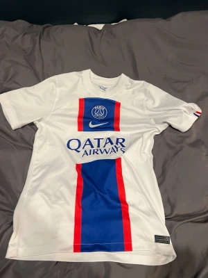 PSG vit matchtröja Nike - Snygg vit PSG matchtröja från Nike med blå och röda detaljer på framsidan. Tröjan har klubbmärke och Nike-logga på bröstet samt Qatar Airways som sponsortryck. Tillverkad i lätt och ventilerande material, perfekt för fotboll eller träning.